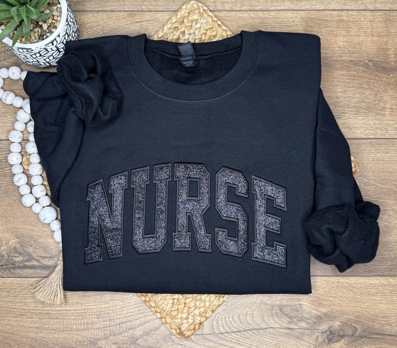 May include: A black crewneck sweatshirt with the word "NURSE" in large, glittery, gray block letters across the chest. The sweatshirt is folded on a woven mat, with a small potted plant and a string of white beads in the background.
