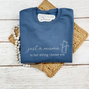 May include: A blue sweatshirt with the text "just a mama in her string cheese era" in white script. A string cheese graphic is on the right. The sweatshirt has a crew neck and is folded on a woven mat.