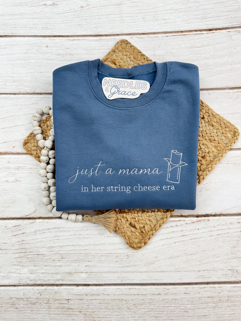 May include: A blue sweatshirt with the text "just a mama in her string cheese era" in white script. A string cheese graphic is on the right. The sweatshirt has a crew neck and is folded on a woven mat.