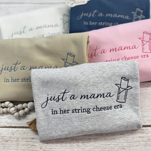 May include: Folded sweatshirts in various colors, including gray, pink, and navy blue, are displayed. Each sweatshirt features the embroidered phrase "just a mama in her string cheese era" with a string cheese graphic.