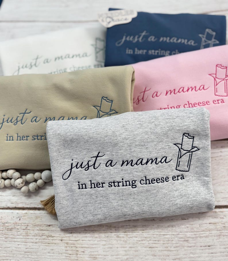 May include: Folded sweatshirts in various colors, including gray, pink, and navy blue, are displayed. Each sweatshirt features the embroidered phrase "just a mama in her string cheese era" with a string cheese graphic.