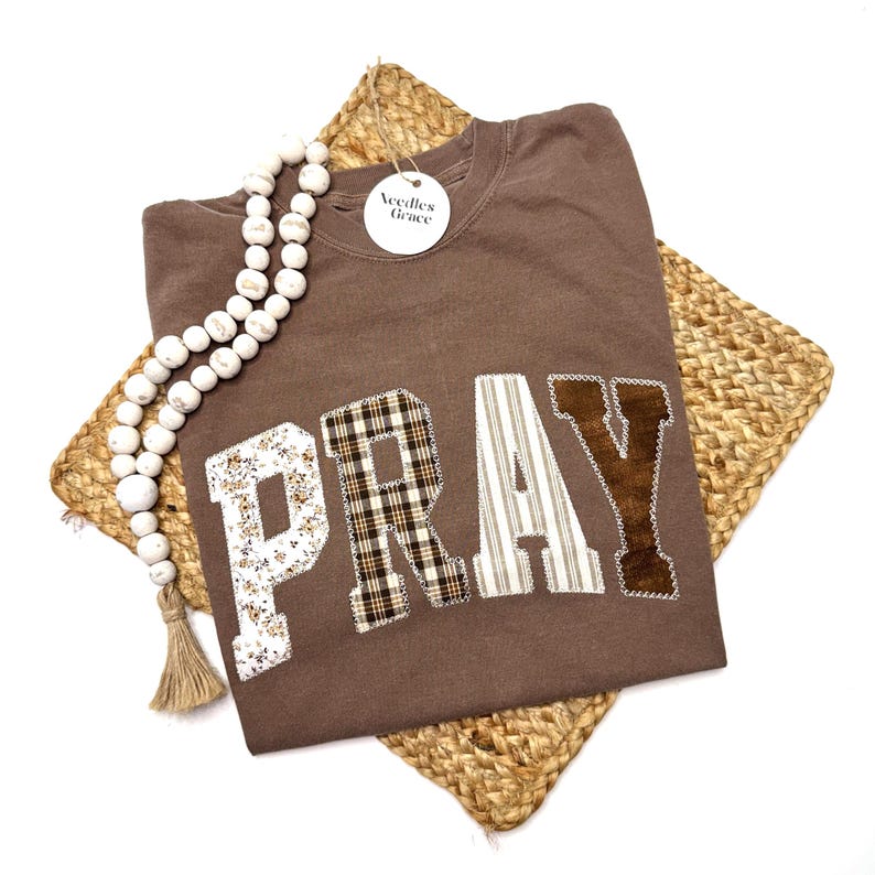 May include: A brown sweatshirt with the word "PRAY" appliqued in various patterned fabrics. The letter "P" has a floral design, "R" has a plaid pattern, "A" has vertical stripes, and "Y" has a brown velvet texture. A white beaded garland and a tag that says "Needles Grace" are also visible.