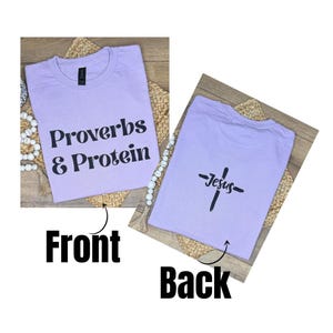 May include: A lilac t-shirt with the text "Proverbs & Protein" on the front. The back of the shirt features a cross with the word "Jesus" underneath. The shirt is displayed on a woven mat.