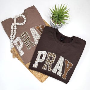 May include: Two brown sweatshirts with the word "PRAY" appliqued on the front. The letters are made of various patterned fabrics, including floral, plaid, and striped designs. A decorative beaded strand and a small potted plant are also in the image.