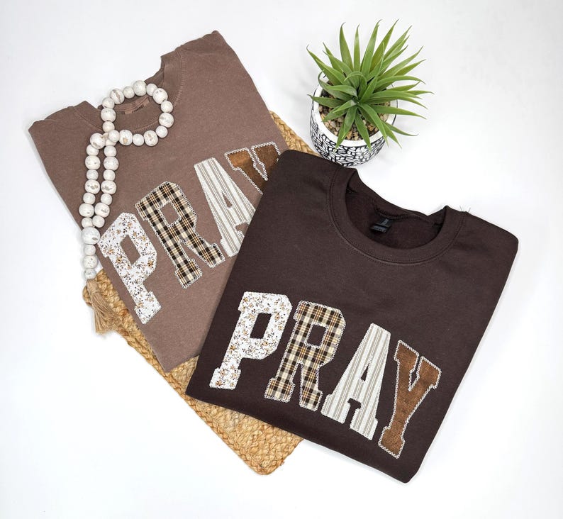 May include: Two brown sweatshirts with the word "PRAY" appliqued on the front. The letters are made of various patterned fabrics, including floral, plaid, and striped designs. A decorative beaded strand and a small potted plant are also in the image.