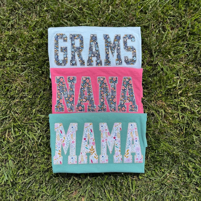 Pu&ograve; includere: Tre magliette piegate in azzurro, rosa e verde acqua, ognuna con una parola a fantasia floreale: "GRAMS", "NANA" e "MAMA". Le magliette sono disposte su erba verde, creando un regalo colorato e personalizzato.