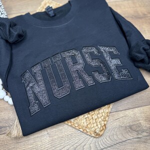 May include: Black crewneck sweatshirt with the word "NURSE" in large, glittery, block letters across the chest. The sweatshirt is folded on a woven, circular placemat, with a wooden surface in the background.