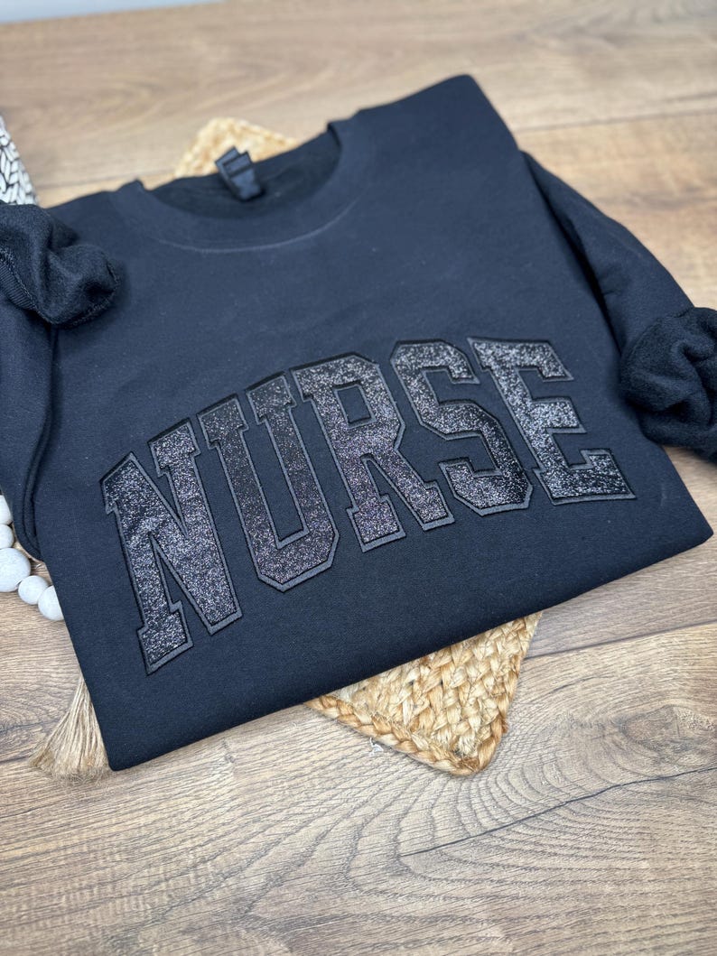 May include: Black crewneck sweatshirt with the word "NURSE" in large, glittery, block letters across the chest. The sweatshirt is folded on a woven, circular placemat, with a wooden surface in the background.
