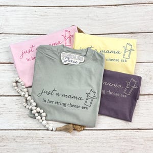 May include: A collection of t-shirts in various colors, including pink, yellow, gray, and purple. Each shirt features the phrase "just a mama in her string cheese era" with a string cheese graphic. A beaded necklace and a tag that says "Needles & Grace" are also visible.