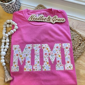 Pu&ograve; includere: Una maglietta rosa acceso con la scritta "MIMI" applicata con un motivo floreale. La maglietta &egrave; piegata su un tappetino intrecciato. Un cartello di legno con la scritta "Needles & Grace" &egrave; sopra.