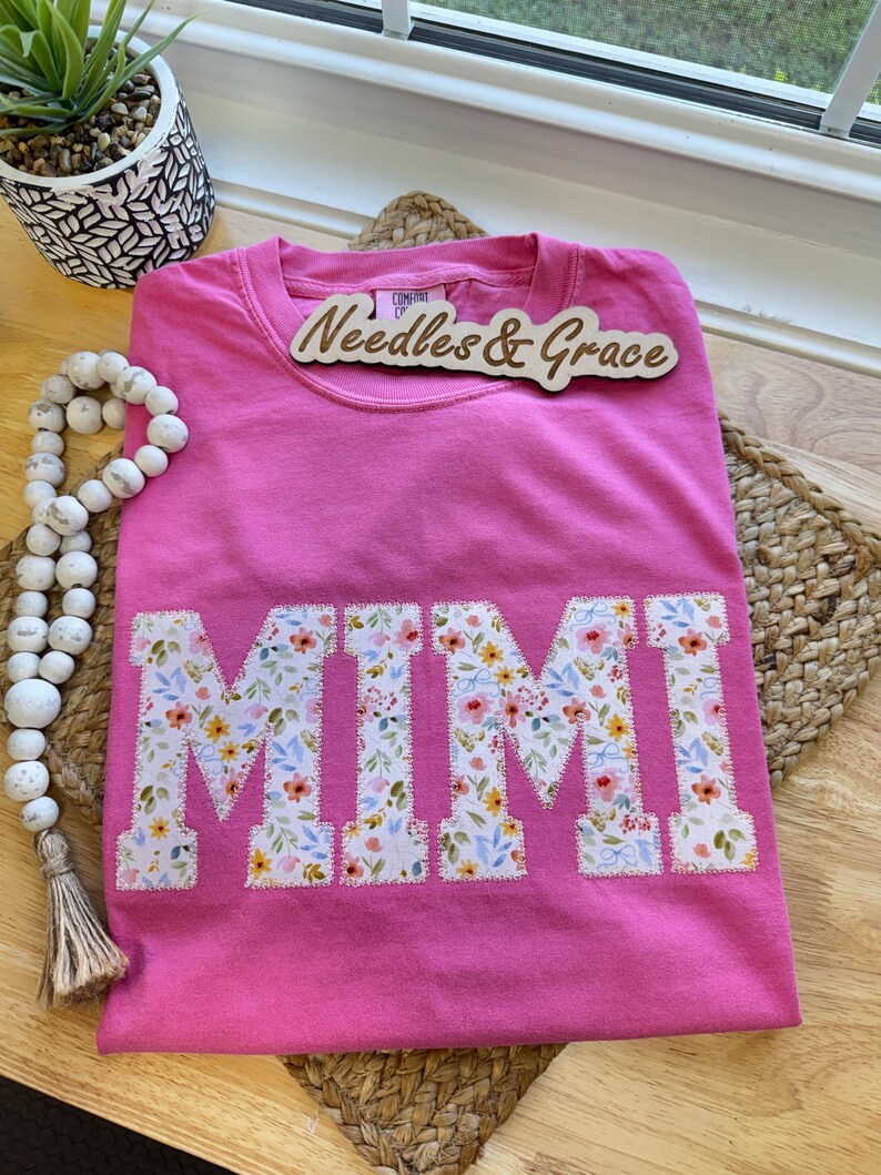 Pu&ograve; includere: Una maglietta rosa acceso con la scritta "MIMI" applicata con un motivo floreale. La maglietta &egrave; piegata su un tappetino intrecciato. Un cartello di legno con la scritta "Needles & Grace" &egrave; sopra.