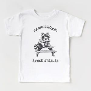 May include: White t-shirt with a black and white illustration of a raccoon sitting at a picnic table and eating a donut. The text "Professional Snack Stealer" is printed below the image.