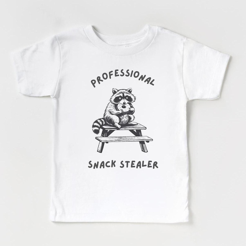 May include: White t-shirt with a black and white illustration of a raccoon sitting at a picnic table and eating a donut. The text "Professional Snack Stealer" is printed below the image.