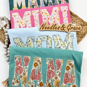 Pu&ograve; includere: Quattro magliette colorate con lettere patchwork floreali che compongono "MAMA", "MIMI", "MOM" e "NANA". Le magliette sono in verde acqua, rosa e azzurro. &Egrave; visibile anche un cartello di legno con la scritta "Needles & Grace".