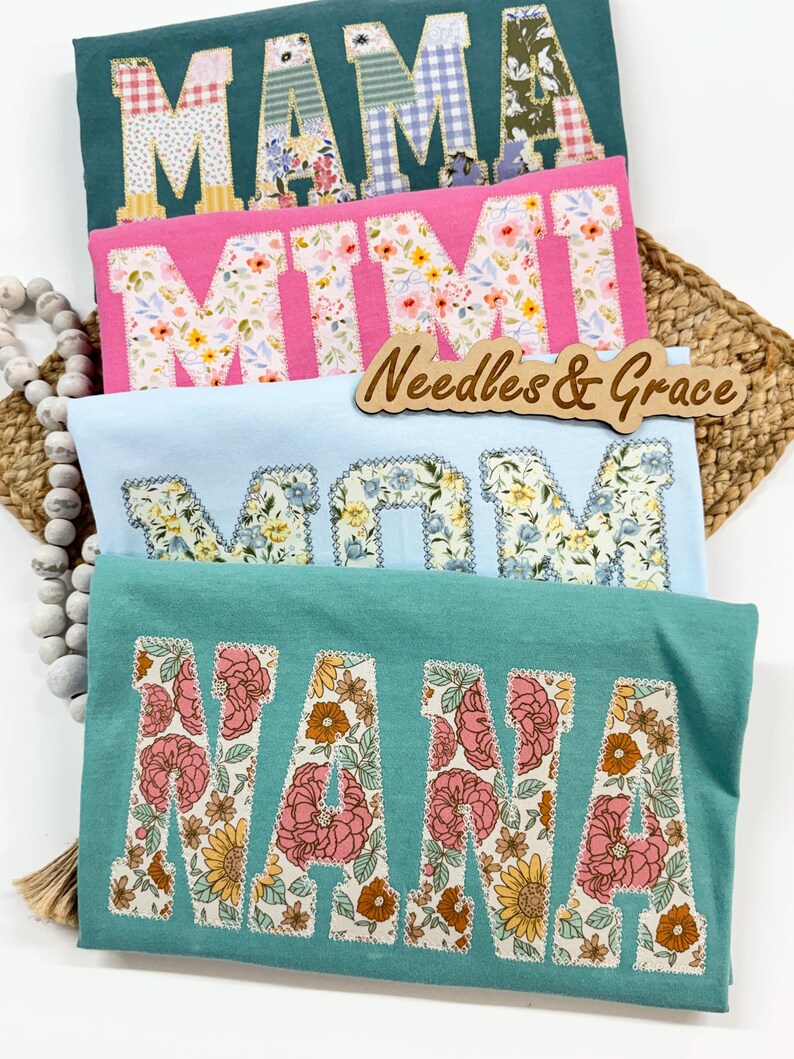 Pu&ograve; includere: Quattro magliette colorate con lettere patchwork floreali che compongono "MAMA", "MIMI", "MOM" e "NANA". Le magliette sono in verde acqua, rosa e azzurro. &Egrave; visibile anche un cartello di legno con la scritta "Needles & Grace".
