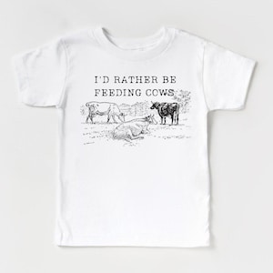 Op de afbeelding: Wit T-shirt met zwarte tekst die "Ik zou liever koeien voeren" luidt. De tekst is gecentreerd op het shirt. Er zijn drie zwart-wit illustraties van koeien in een weiland onder de tekst.