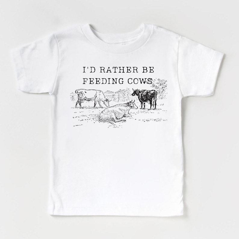 Op de afbeelding: Wit T-shirt met zwarte tekst die "Ik zou liever koeien voeren" luidt. De tekst is gecentreerd op het shirt. Er zijn drie zwart-wit illustraties van koeien in een weiland onder de tekst.