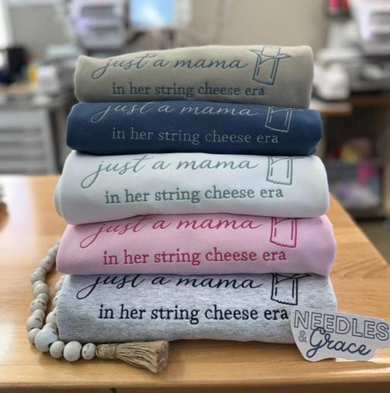 May include: A stack of five folded sweatshirts in various colors: tan, navy blue, white, pink, and gray. Each sweatshirt has the embroidered text "just a mama in her string cheese era". A sticker with the text "Needles & Grace" is visible.