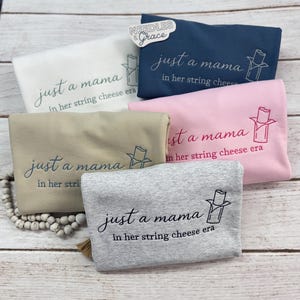May include: Five folded sweatshirts in various colors: white, navy blue, pink, tan, and gray. Each sweatshirt has the embroidered text "just a mama in her string cheese era" with a string cheese graphic.
