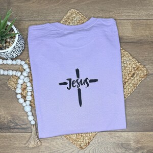 May include: A lilac t-shirt featuring a black cross and the word "Jesus". The shirt is placed on a woven mat. A small potted plant and decorative beads are visible in the background.