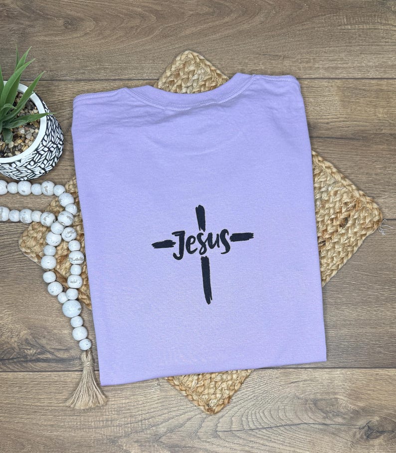 May include: A lilac t-shirt featuring a black cross and the word "Jesus". The shirt is placed on a woven mat. A small potted plant and decorative beads are visible in the background.