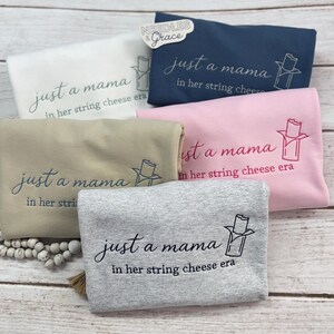 May include: A collection of folded sweatshirts in various colors: white, navy blue, pink, tan, and gray. Each sweatshirt features the phrase "just a mama in her string cheese era" in cursive script, along with a string cheese graphic.