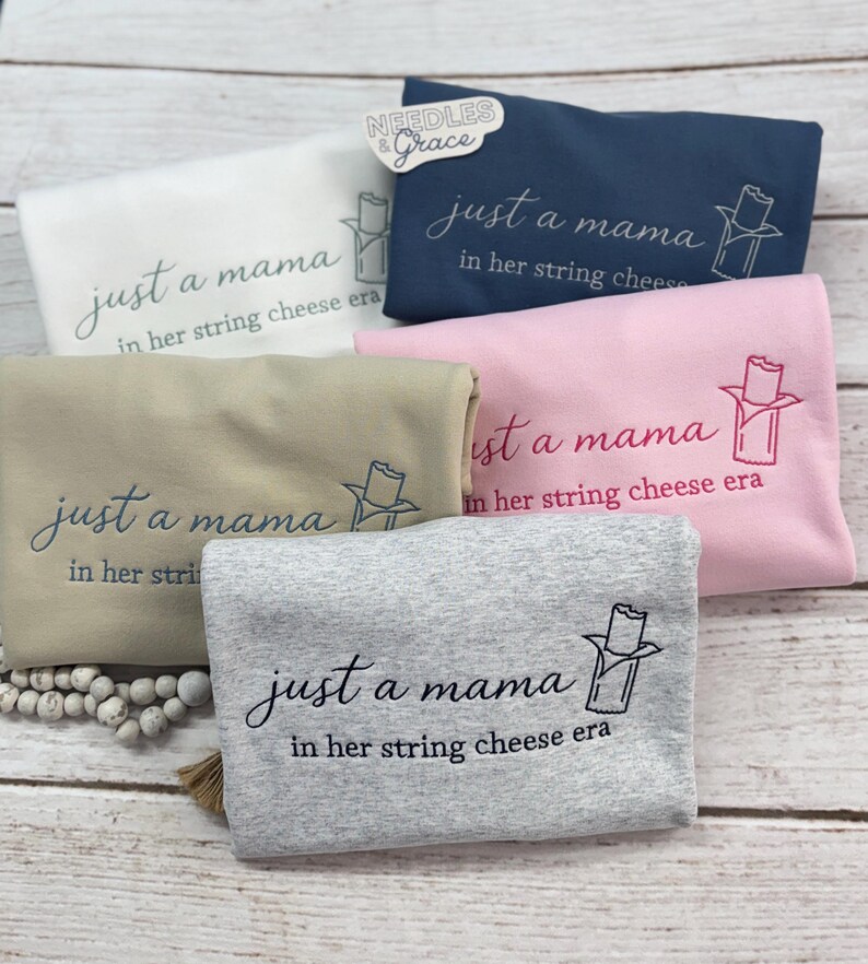 May include: A collection of folded sweatshirts in various colors: white, navy blue, pink, tan, and gray. Each sweatshirt features the phrase "just a mama in her string cheese era" in cursive script, along with a string cheese graphic.