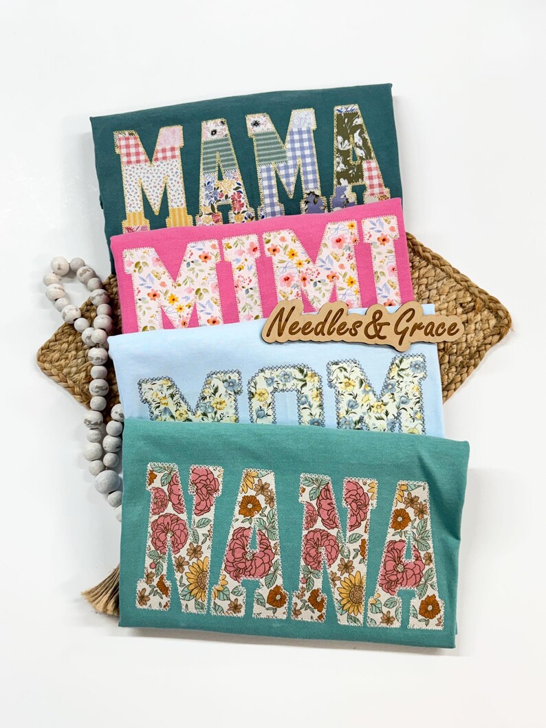 Pu&ograve; includere: Quattro magliette in verde acqua, rosa e azzurro, ognuna con un nome diverso in lettere patchwork floreali: MAMA, MIMI, MOM e NANA. &Egrave; visibile anche un cartello di legno con la scritta "Needles & Grace".