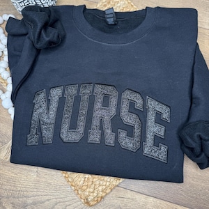 May include: A black sweatshirt with the word "NURSE" in large, glittery, block letters across the chest. The sweatshirt is folded, with the sleeves visible. A small plant in a white and black patterned pot is in the background.