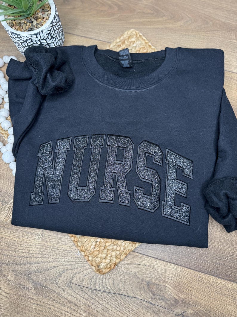 May include: A black sweatshirt with the word "NURSE" in large, glittery, block letters across the chest. The sweatshirt is folded, with the sleeves visible. A small plant in a white and black patterned pot is in the background.