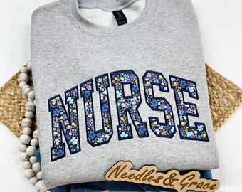 May include: A gray sweatshirt with the word "NURSE" in navy blue floral letters. The sweatshirt is folded over blue jeans. A wooden sign that says "Needles & Grace" is on top of the jeans. A beaded garland and woven mat are also in the image.