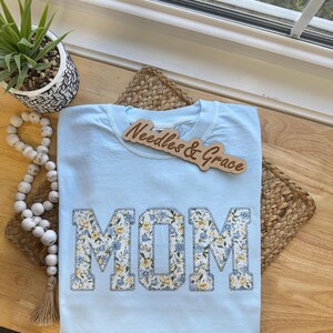 Pu&ograve; includere: Una maglietta azzurra con la scritta "MOM" in lettere patchwork floreali. La maglietta ha un girocollo e maniche corte. Un cartello di legno con la scritta "Needles & Grace" &egrave; sopra la maglietta.