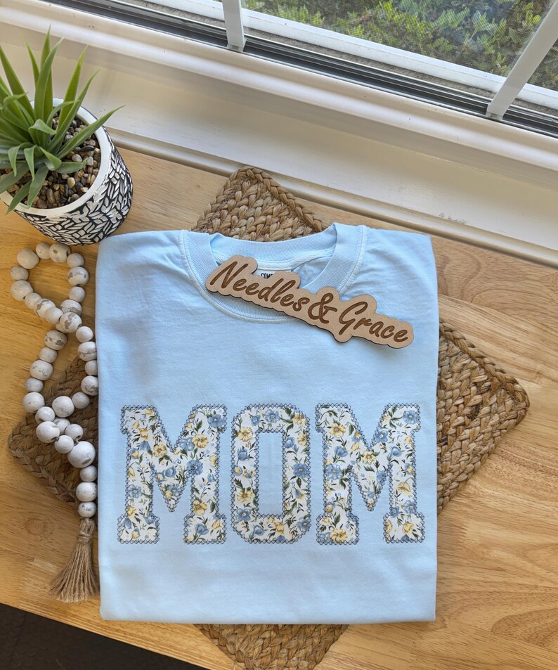 Pu&ograve; includere: Una maglietta azzurra con la scritta "MOM" in lettere patchwork floreali. La maglietta ha un girocollo e maniche corte. Un cartello di legno con la scritta "Needles & Grace" &egrave; sopra la maglietta.