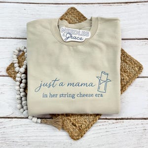 May include: Beige crewneck sweatshirt with the text "just a mama in her string cheese era" in blue script. A blue graphic of a string cheese stick is next to the text. The sweatshirt is on a woven mat with a beaded garland.