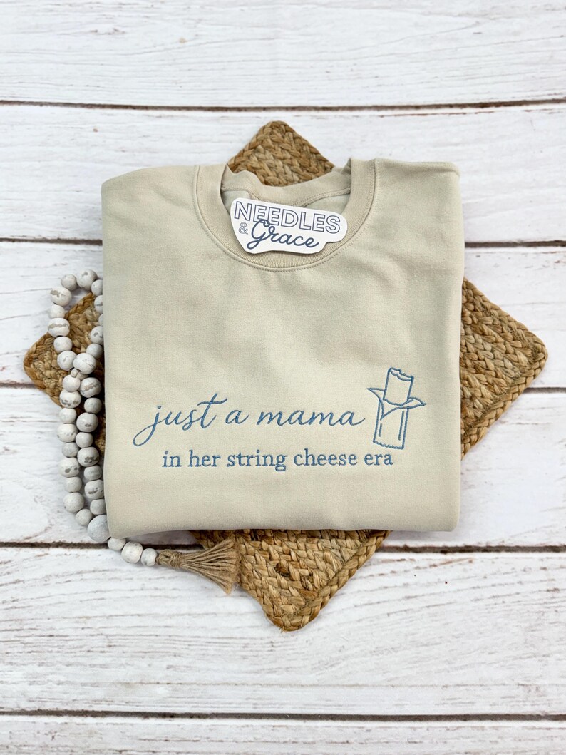 May include: Beige crewneck sweatshirt with the text "just a mama in her string cheese era" in blue script. A blue graphic of a string cheese stick is next to the text. The sweatshirt is on a woven mat with a beaded garland.