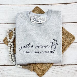 May include: A heather gray sweatshirt with the text "just a mama in her string cheese era" and a string cheese graphic. The sweatshirt is folded on a woven placemat and a strand of white beads.