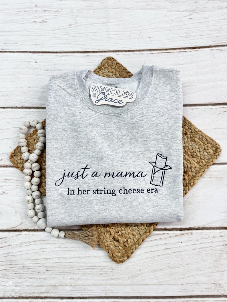 May include: A heather gray sweatshirt with the text "just a mama in her string cheese era" and a string cheese graphic. The sweatshirt is folded on a woven placemat and a strand of white beads.