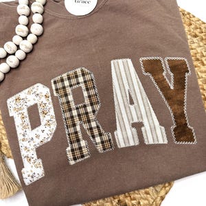 May include: A brown long-sleeve shirt with the word "PRAY" appliqued on the front. The letters are made of various patterned fabrics, including floral, plaid, and striped designs. A tag that says "Needles Grace" is attached to the shirt.