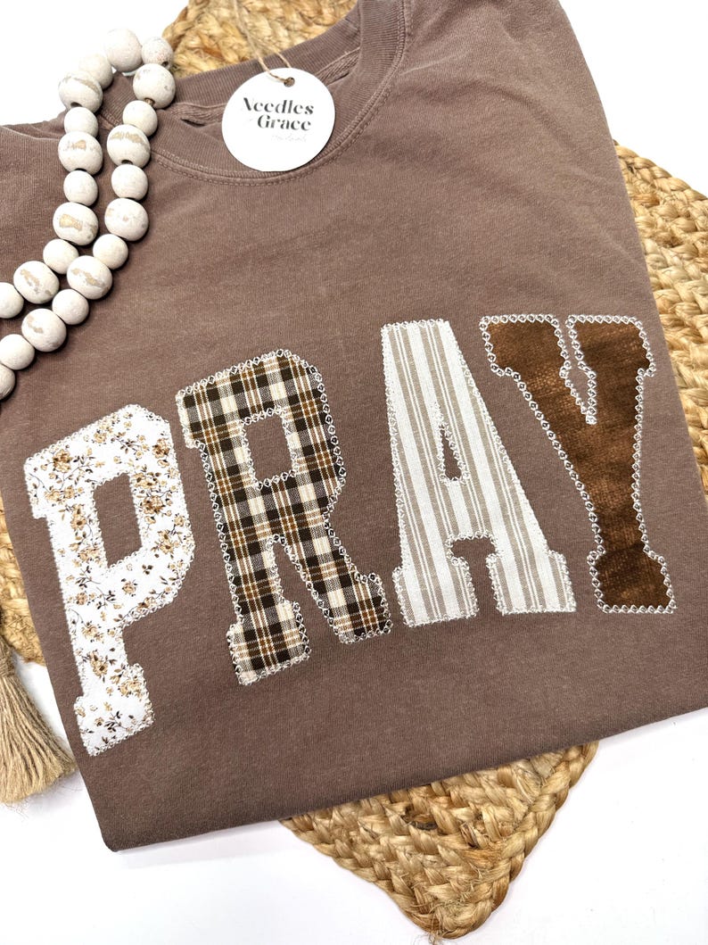 May include: A brown long-sleeve shirt with the word "PRAY" appliqued on the front. The letters are made of various patterned fabrics, including floral, plaid, and striped designs. A tag that says "Needles Grace" is attached to the shirt.