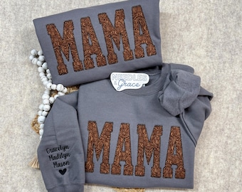 Könnte beinhalten: Zwei graue Sweatshirts mit dem Wort "MAMA" in brauner, strukturierter Schrift. Eines ist gefaltet, das andere hat Namen auf dem Ärmel. Die Sweatshirts sind von "Needles & Grace".