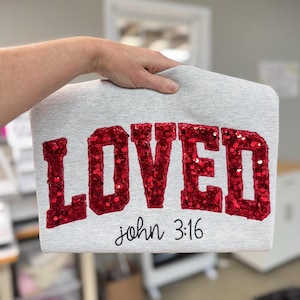 May include: A gray sweatshirt with the word "LOVED" in large, red sequined letters. Below the word is the text "john 3:16" in a simple black font. The sweatshirt is held up by a hand.