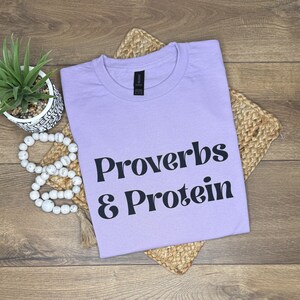 May include: A lilac t-shirt featuring the text "Proverbs & Protein" in black lettering. The shirt is placed on a woven mat, with a small potted plant and decorative beads. The t-shirt has a round neck.