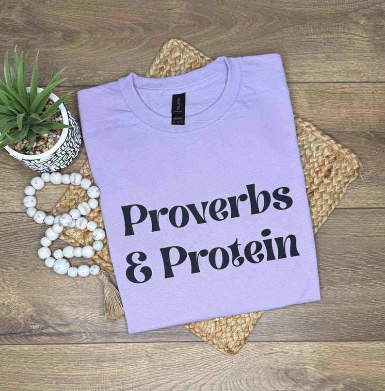 May include: A lilac t-shirt featuring the text "Proverbs & Protein" in black lettering. The shirt is placed on a woven mat, with a small potted plant and decorative beads. The t-shirt has a round neck.