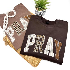 May include: Two brown sweatshirts with the word "PRAY" appliqued across the front in various patterned fabrics. One is a lighter brown, the other a darker brown. A white beaded necklace and a small potted plant are also visible.