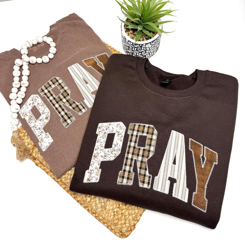 May include: Two brown sweatshirts with the word "PRAY" appliqued across the front in various patterned fabrics. One is a lighter brown, the other a darker brown. A white beaded necklace and a small potted plant are also visible.
