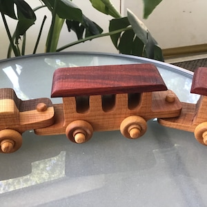 May include: A wooden toy train set with three cars. The engine car is made of light wood with a dark wood stripe. The middle car has a red wood roof and three windows. The last car is made of light wood with a red wood roof.