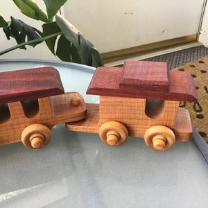 May include: A wooden toy train with two cars. The first car has a red roof and the second car has a brown roof. Both cars have wooden wheels.