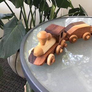 May include: A wooden toy train set with a locomotive and two cars. The train is made of natural wood with a light brown finish. The locomotive has a brown roof and a black chimney. The cars are both brown with a light brown stripe.