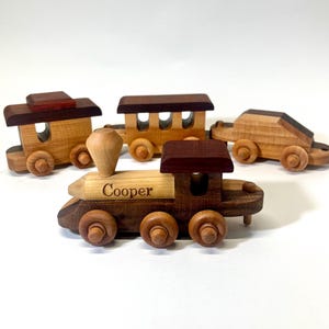May include: A wooden toy train set with a locomotive, a passenger car, and a car. The locomotive has a brown and red roof and the name "Cooper" engraved on the side. The passenger car has a brown roof and three windows. The car is brown and has a black stripe.