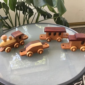 May include: A set of four wooden toy train cars, including an engine, a passenger car, and two boxcars. The cars are made of different types of wood, giving them a unique look. The cars are painted in natural wood tones and have black wheels.